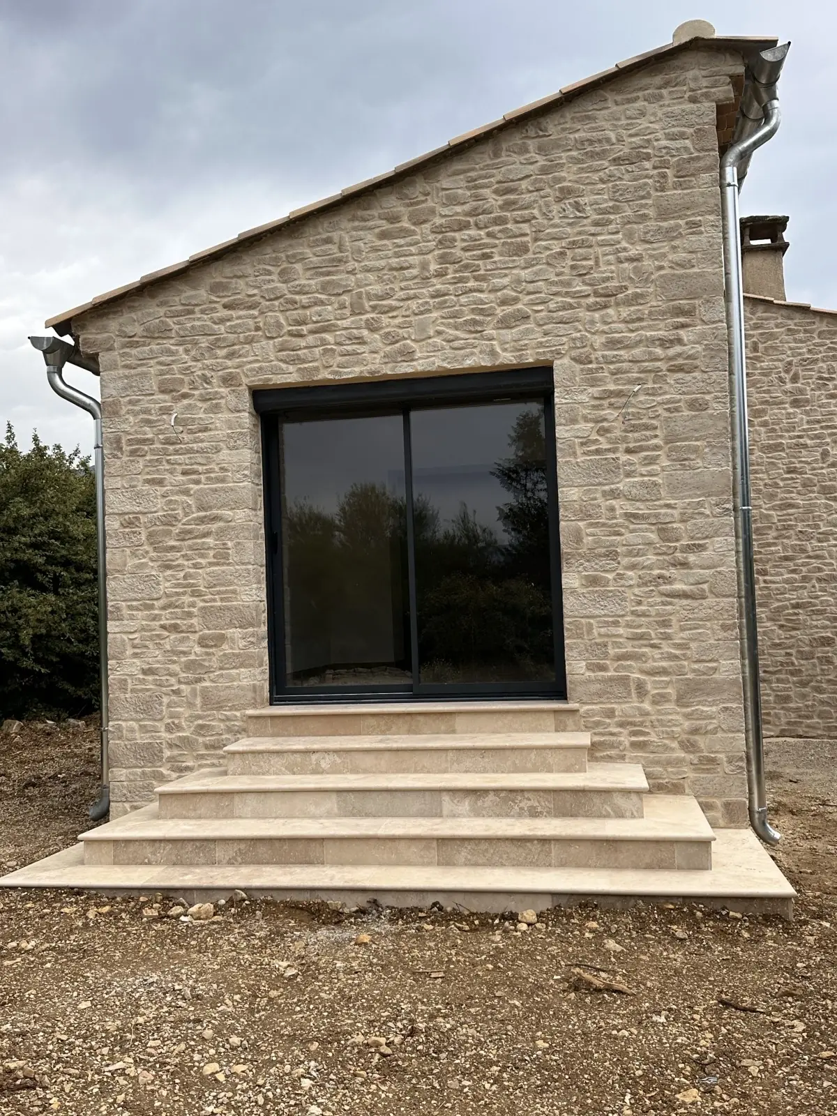 Petit bâtiment en pierre de parement, grande baie vitrée noire, escalier d'entrée en pierre claire