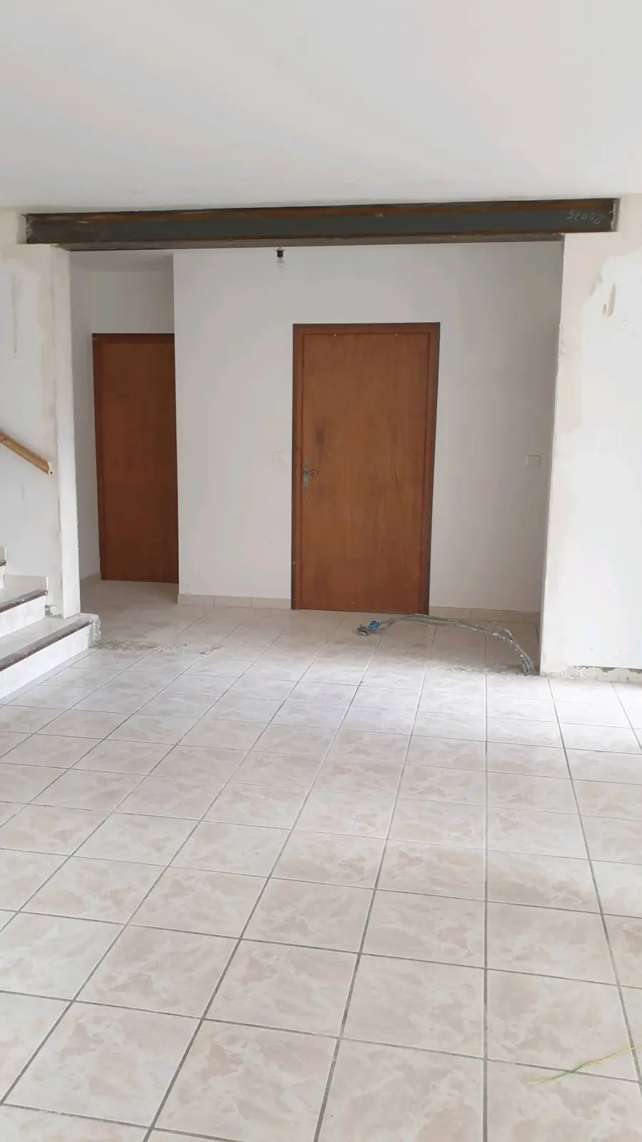 Pièce au carrelage clair, murs blancs, deux portes en bois, poutre au plafond