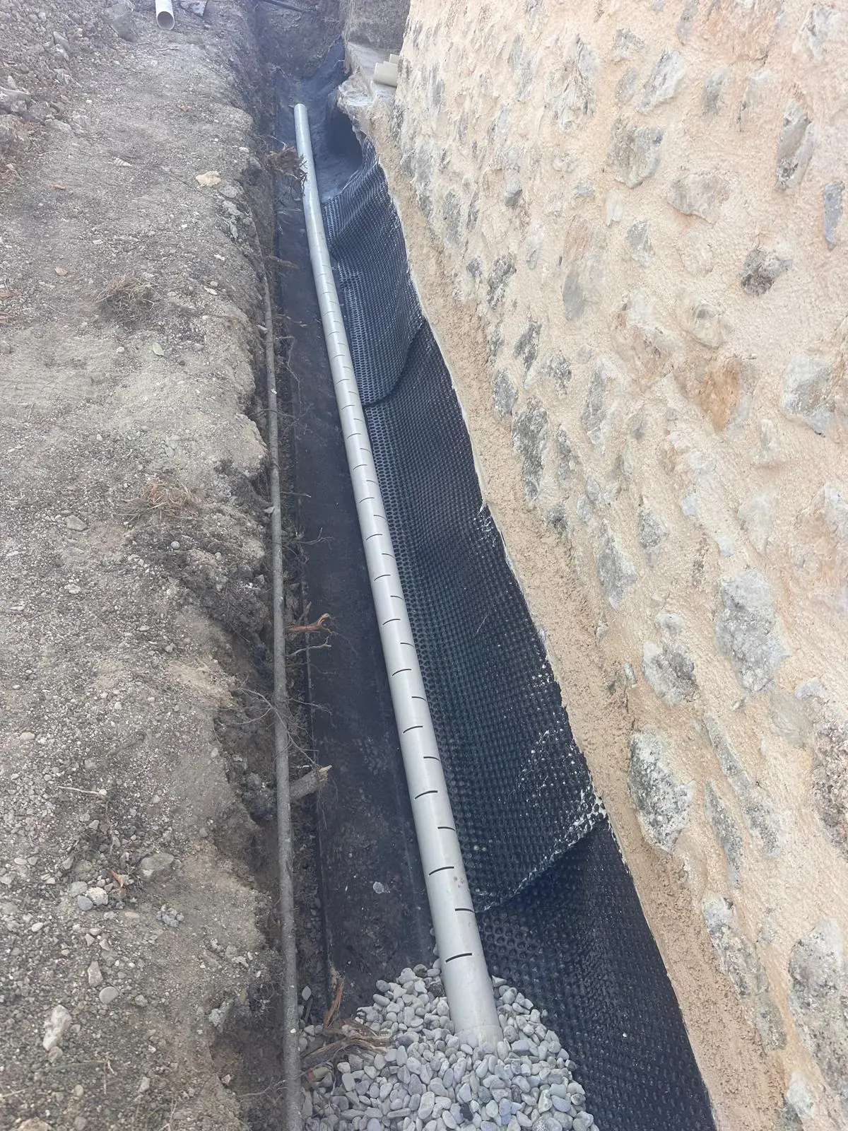 Drain posé sur graviers au pied d’un mur en pierre, nappe noire contre le mur