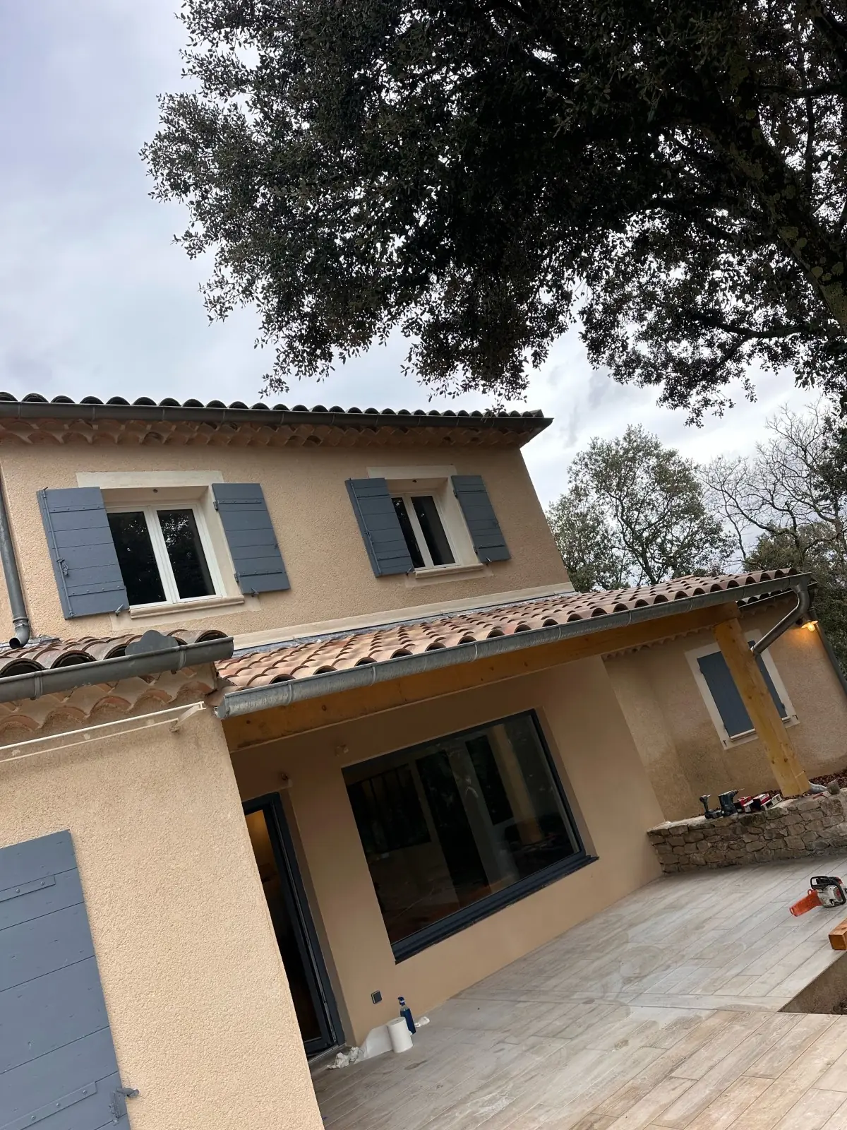 Maison à étage enduite, volets bleus : avancée de toit en tuiles au-dessus d'une terrasse et d'une grande baie, sous un chêne