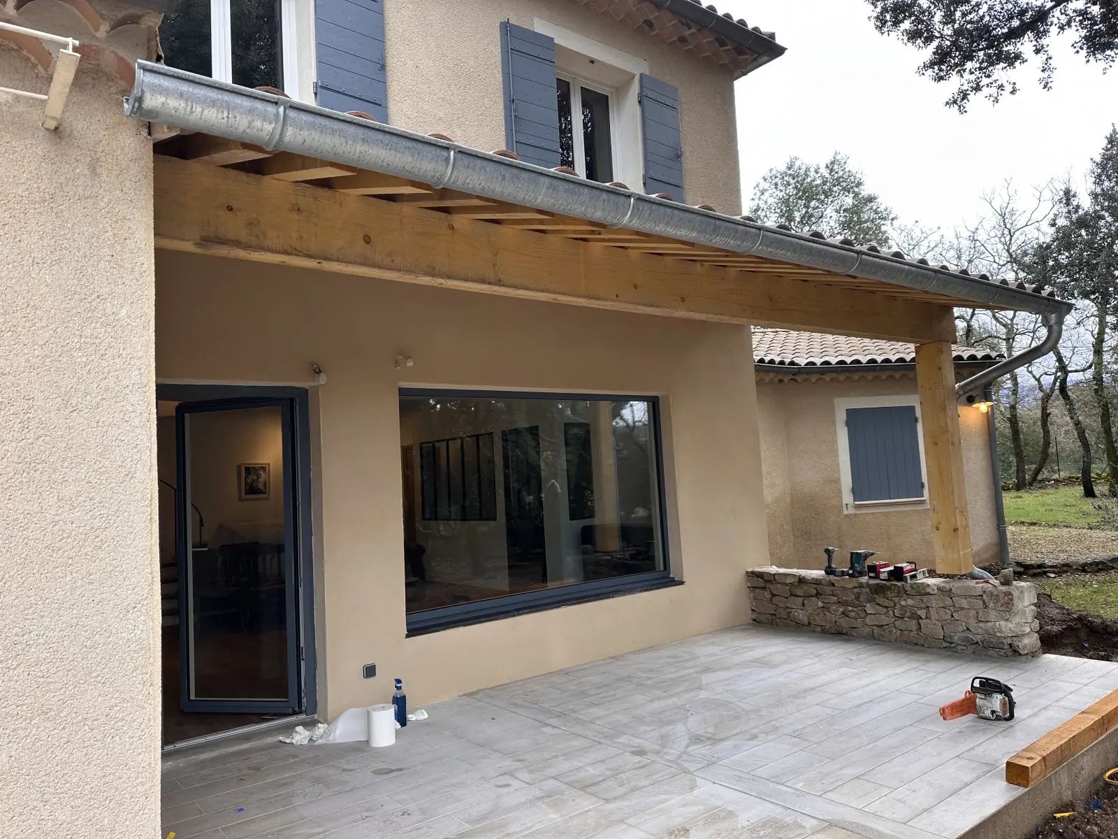 Avancée de toit en tuiles sur poteau bois au-dessus d'une terrasse dallée, devant une grande baie