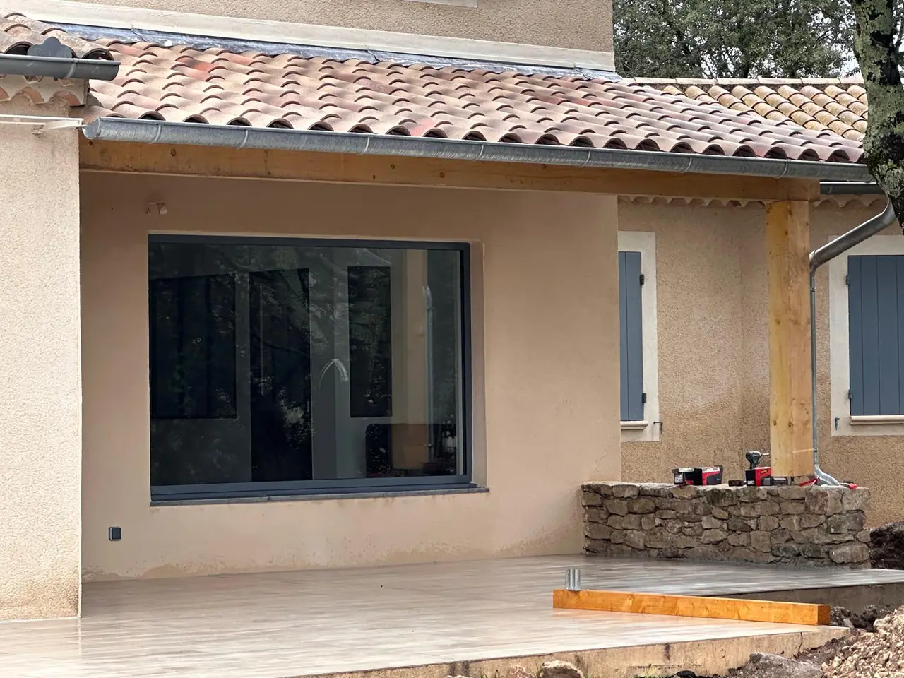 Façade sous une avancée de toit en tuiles : grande baie, terrasse carrelée, muret en pierre