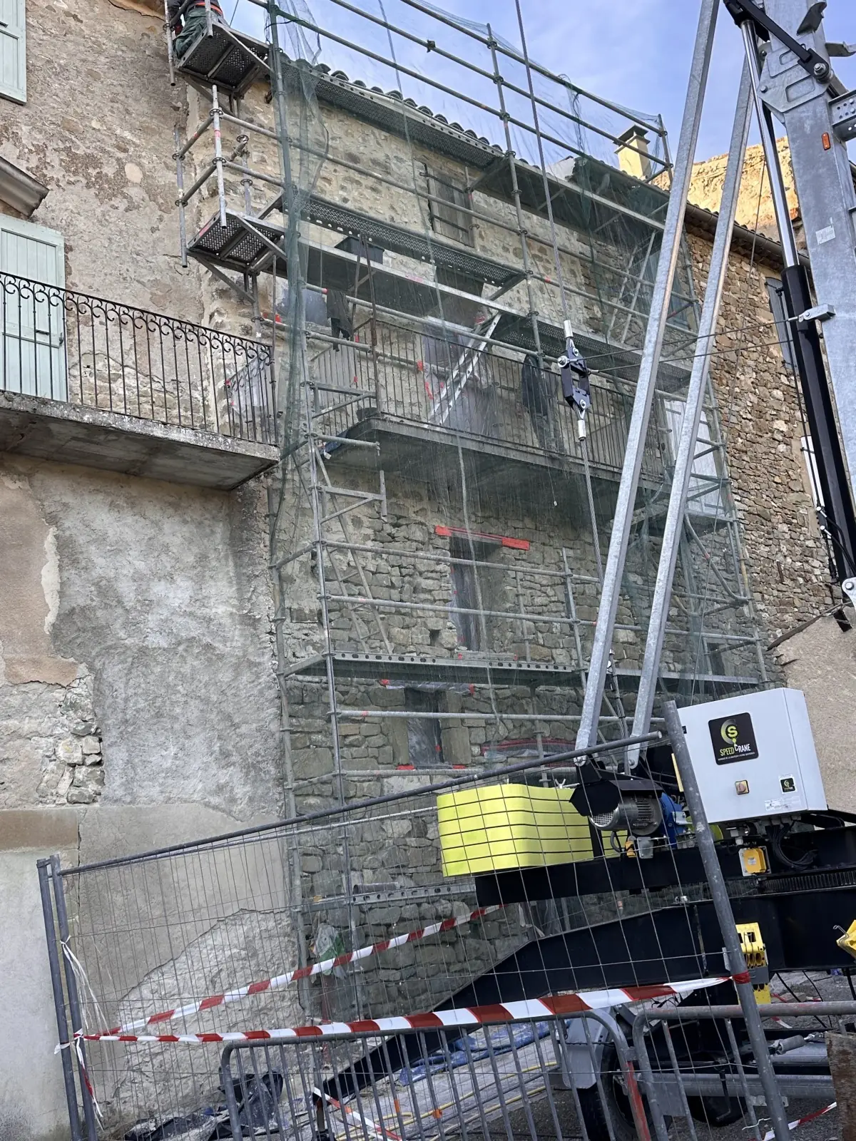 Immeuble en pierre de bourg sous échafaudage, balcon en fer forgé, chantier balisé dans la rue