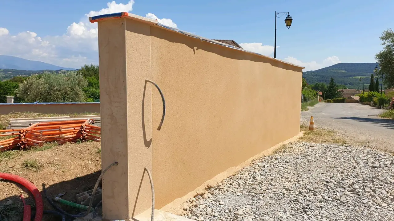 Mur de clôture enduit ocre avec son chaperon, gravier au pied, collines au fond
