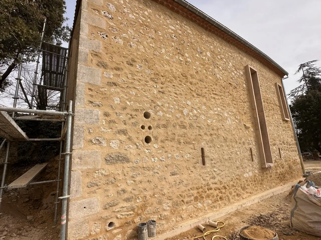 Mur latéral en pierre rejointoyé, longue fenêtre étroite à encadrement bois, échafaudage à gauche