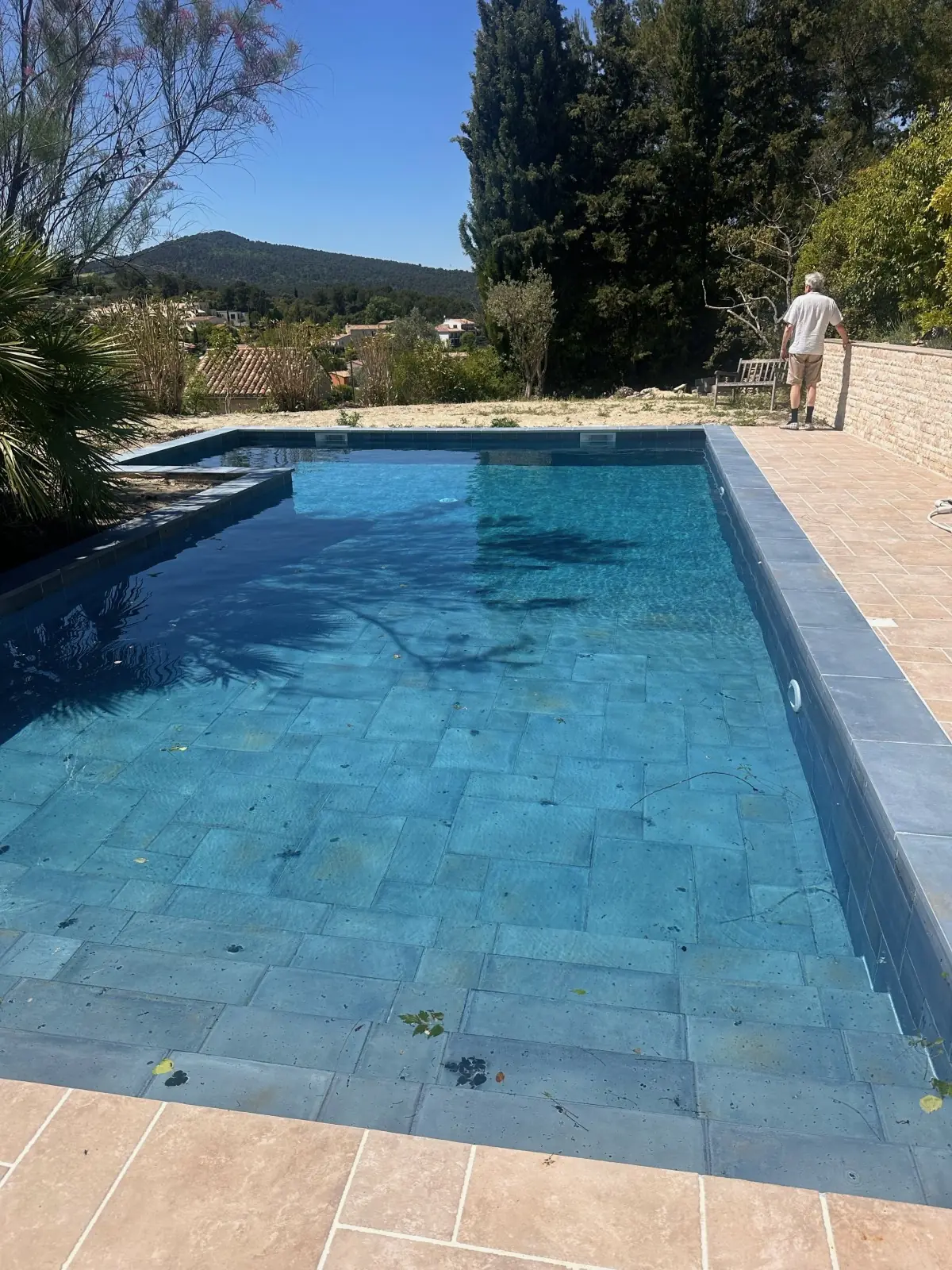 Piscine carrelée en mosaïque bleu-vert, plage en carreaux terre cuite, collines boisées au fond