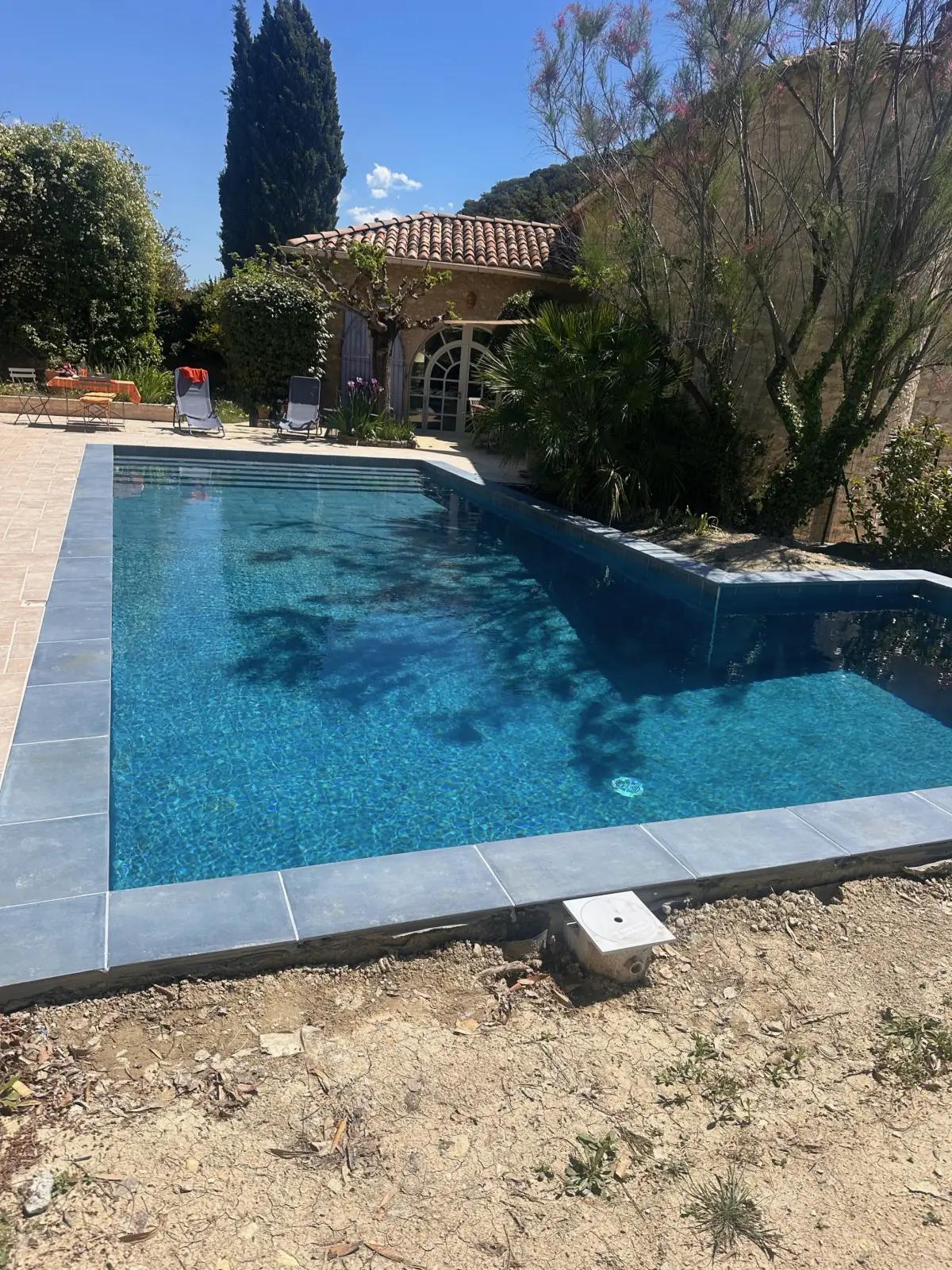 Piscine remplie d’eau bleue devant une maison provençale aux tuiles roses, cyprès, abords encore en terre