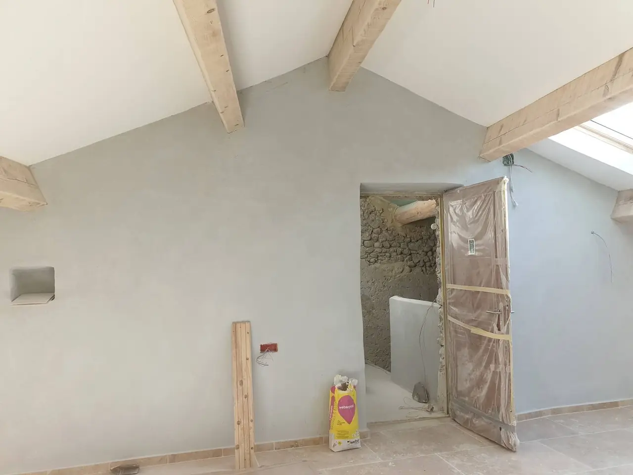 Pièce sous rampant aux murs enduits gris clair, poutres apparentes, fenêtre de toit, porte protégée