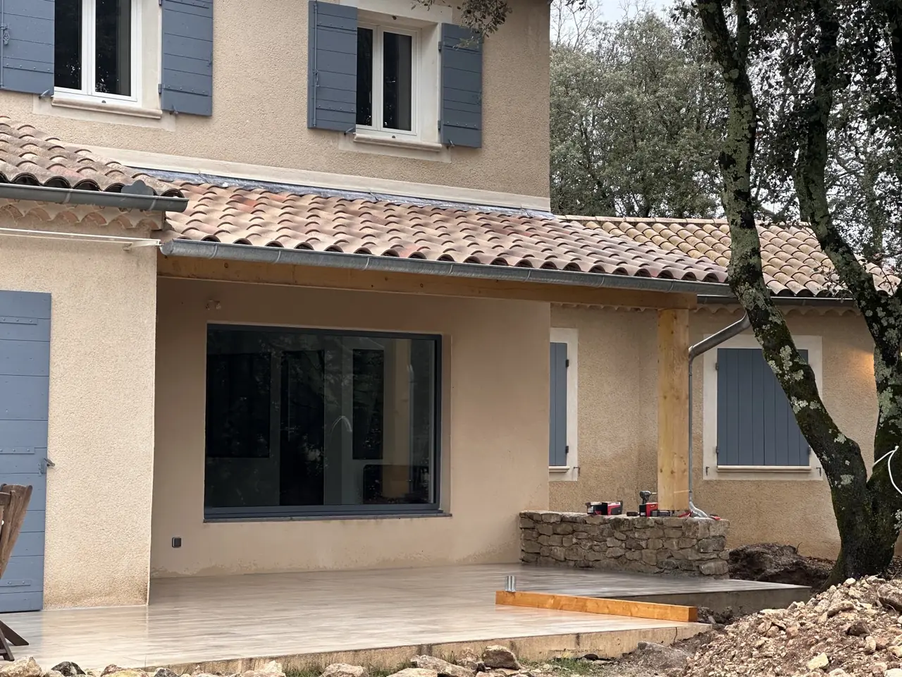 Arrière d’une maison à volets bleus : avancée de toit en tuiles sur poteau bois, chevrons apparents, terrasse carrelée devant une grande baie